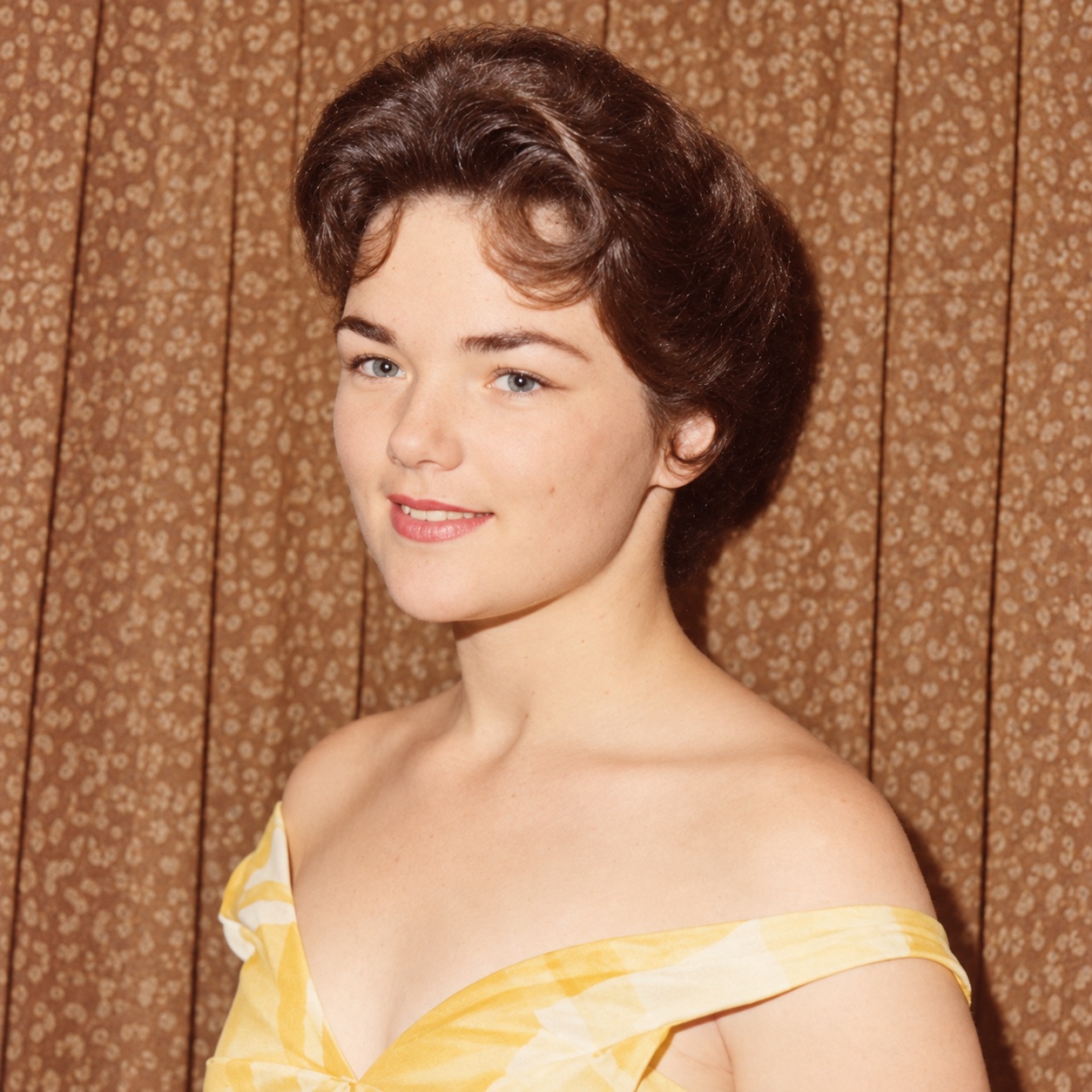 Young Judith wearing an off-the-shoulder yellow dress in front of patterned curtains.